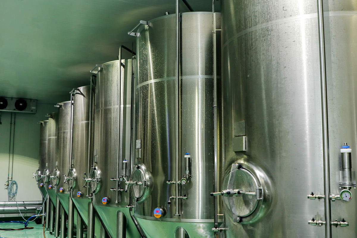 Large stainless steel fermentation tanks lined up in a brewery or industrial facility, with valves and gauges visible on the front of each tank, under bright lighting.