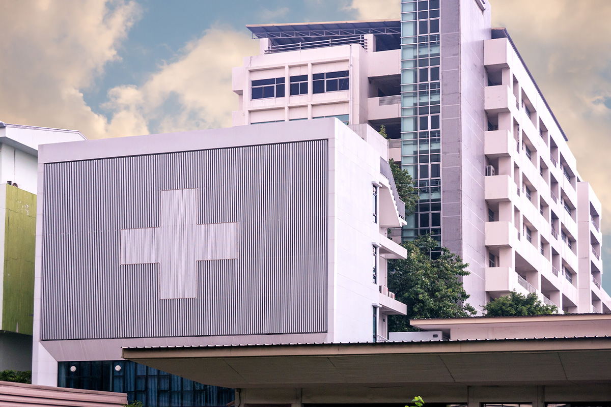A modern hospital building with a large white cross symbol on the facade, multiple floors, glass windows, and a partly cloudy sky in the background.