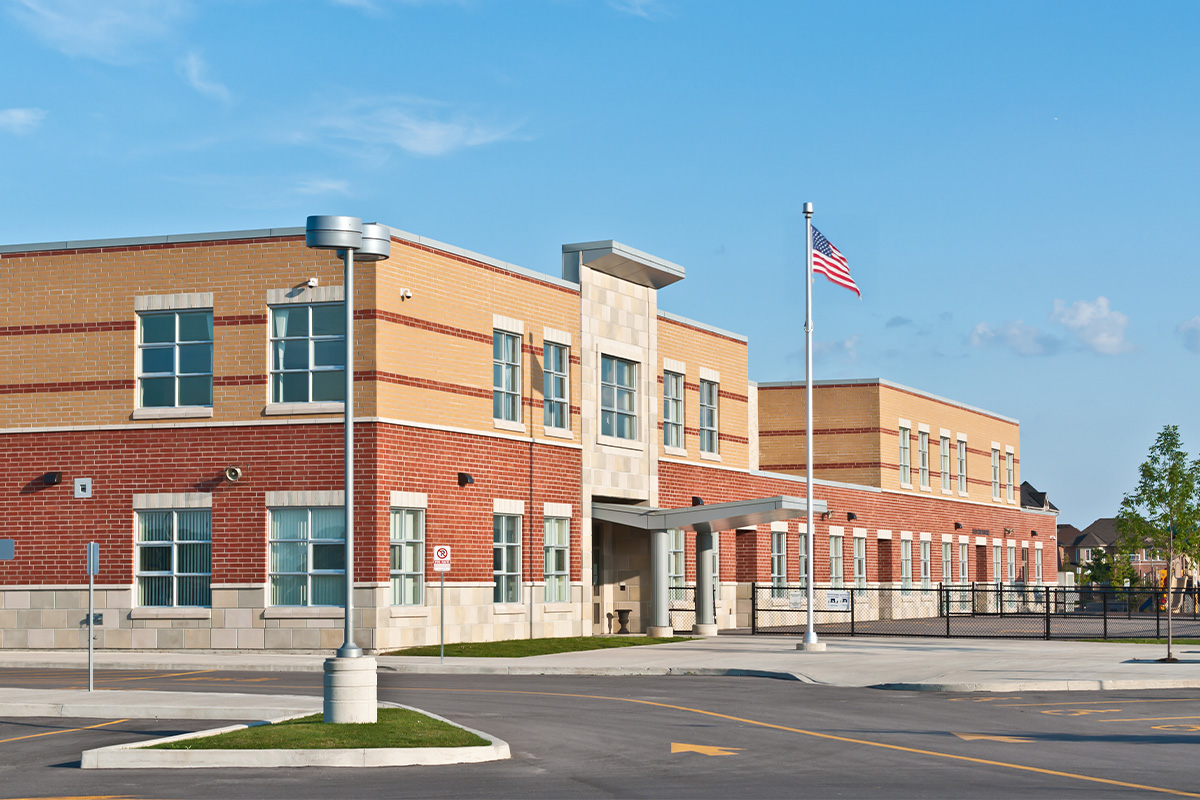 A modern two-story brick school building with large windows, an entrance canopy, and an American flag flying on a pole in front, under a clear blue sky. There is a parking lot and drive-up area in the foreground.